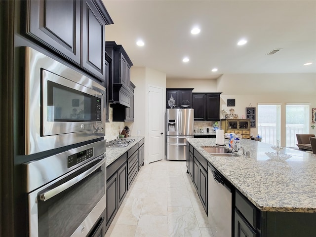 kitchen featuring sink, a kitchen island with sink, stainless steel appliances, light stone countertops, and decorative backsplash