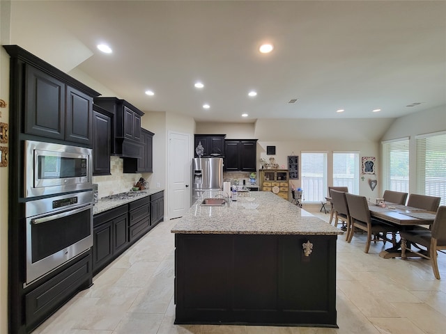 kitchen featuring sink, a center island with sink, stainless steel appliances, light stone countertops, and decorative backsplash