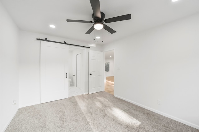 unfurnished bedroom with a closet, light colored carpet, a barn door, connected bathroom, and ceiling fan