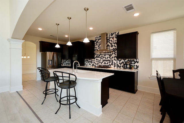 kitchen with pendant lighting, a kitchen island with sink, backsplash, wall chimney range hood, and appliances with stainless steel finishes