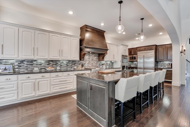 kitchen featuring custom exhaust hood, a large island with sink, white cabinetry, built in appliances, and dark stone counters