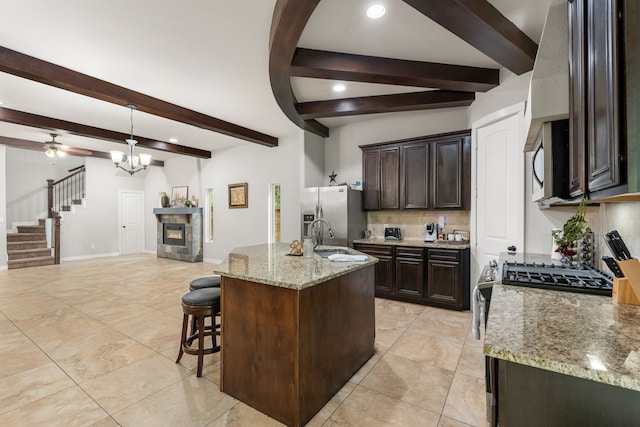 kitchen featuring an island with sink, ceiling fan with notable chandelier, light stone countertops, stainless steel appliances, and beam ceiling