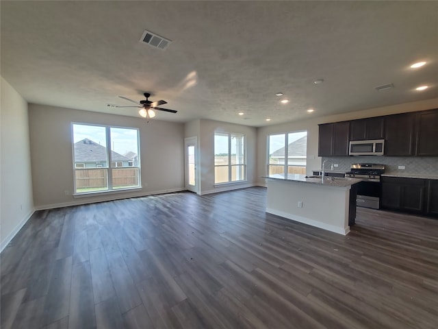 kitchen with ceiling fan, dark hardwood / wood-style flooring, backsplash, an island with sink, and appliances with stainless steel finishes