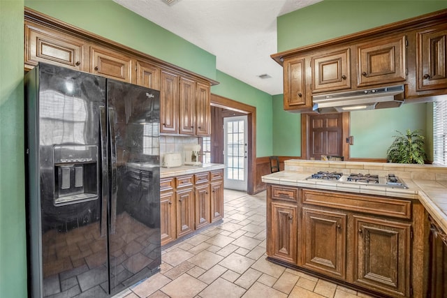 kitchen featuring black refrigerator with ice dispenser, a textured ceiling, tasteful backsplash, tile counters, and stainless steel gas cooktop