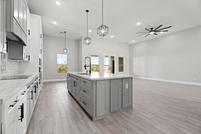 kitchen featuring black electric stovetop, an island with sink, hanging light fixtures, and light wood-type flooring