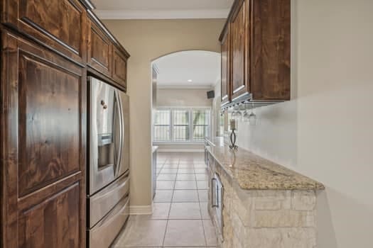 kitchen with dark brown cabinetry, stainless steel fridge, light tile patterned floors, light stone countertops, and crown molding