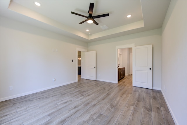 unfurnished bedroom featuring ceiling fan, light hardwood / wood-style flooring, ensuite bath, and a tray ceiling