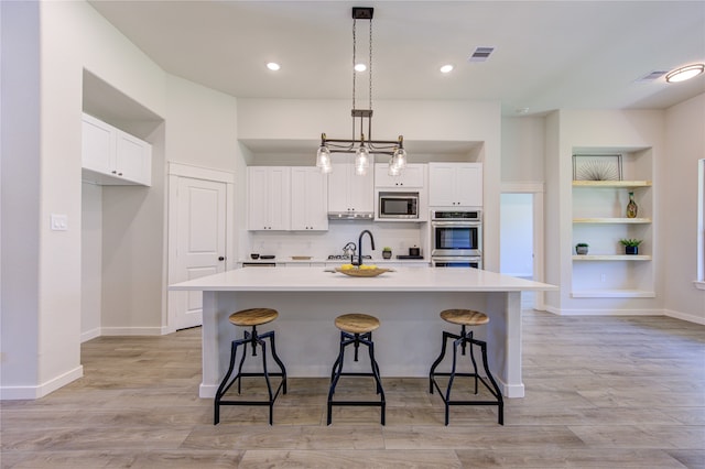 kitchen with appliances with stainless steel finishes, light hardwood / wood-style floors, an island with sink, white cabinets, and pendant lighting