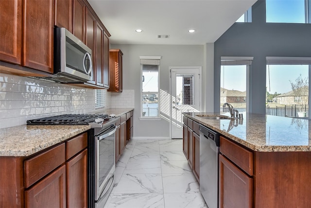 kitchen with an island with sink, light stone countertops, decorative backsplash, stainless steel appliances, and sink