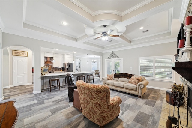 living room featuring ceiling fan with notable chandelier, sink, crown molding, a tiled fireplace, and coffered ceiling