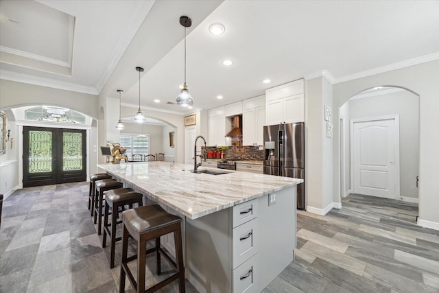 kitchen featuring wall chimney exhaust hood, white cabinetry, an island with sink, hanging light fixtures, and stainless steel fridge