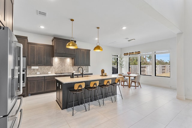 kitchen featuring a kitchen breakfast bar, dark brown cabinets, stainless steel appliances, hanging light fixtures, and an island with sink