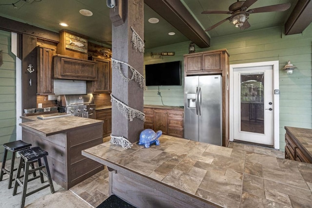 kitchen with wood walls, butcher block countertops, beamed ceiling, ceiling fan, and stainless steel refrigerator with ice dispenser