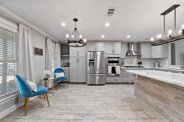 kitchen with wall chimney range hood, hanging light fixtures, gray cabinetry, appliances with stainless steel finishes, and light stone counters