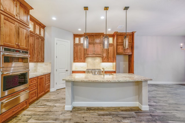 kitchen with light wood-type flooring, an island with sink, stainless steel appliances, decorative light fixtures, and light stone countertops