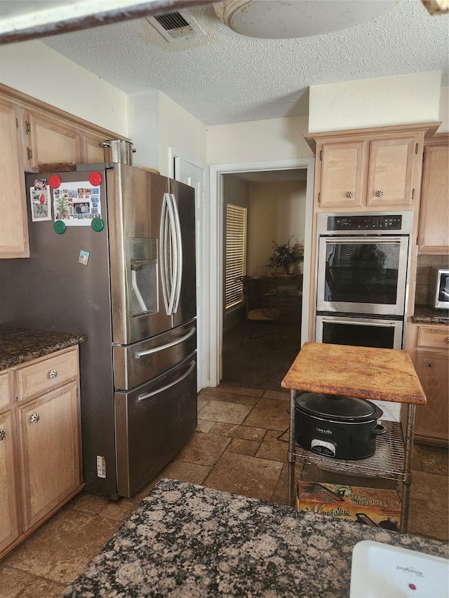 kitchen with light brown cabinets, a textured ceiling, stainless steel appliances, and dark stone counters