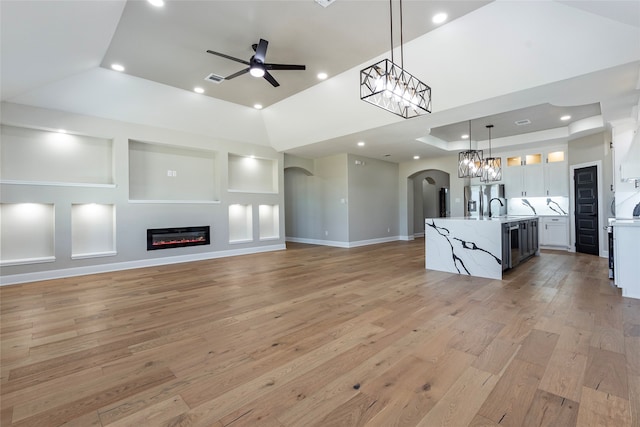 unfurnished living room with a towering ceiling, ceiling fan with notable chandelier, a raised ceiling, light wood-type flooring, and sink