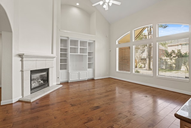 unfurnished living room with high vaulted ceiling, a tiled fireplace, ceiling fan, built in shelves, and dark hardwood / wood-style floors