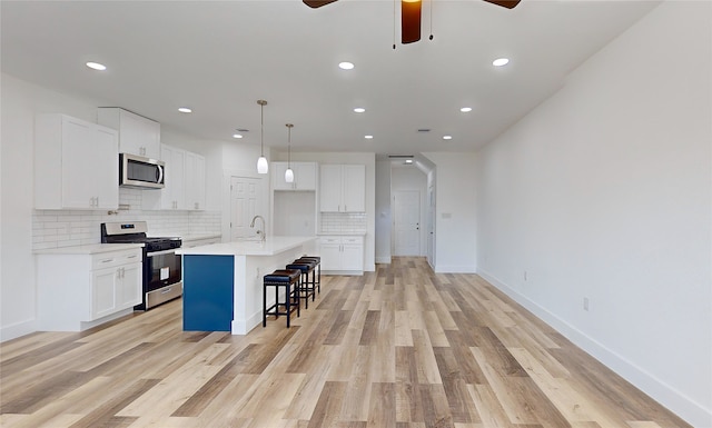 kitchen with light wood-type flooring, a kitchen island with sink, white cabinets, pendant lighting, and appliances with stainless steel finishes