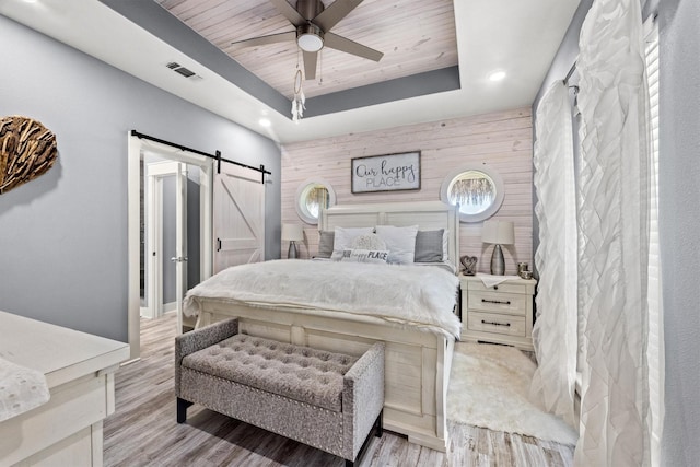 bedroom with wooden walls, ceiling fan, light hardwood / wood-style floors, a raised ceiling, and a barn door