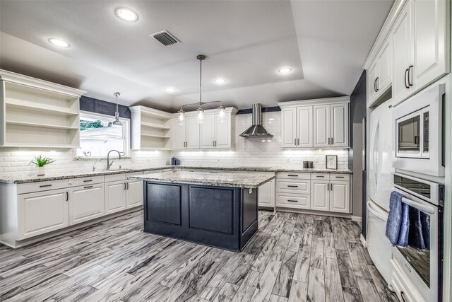 kitchen featuring hanging light fixtures, a kitchen island, wall chimney exhaust hood, oven, and white cabinetry