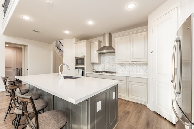 kitchen with wall chimney range hood, a center island with sink, appliances with stainless steel finishes, light hardwood / wood-style floors, and sink