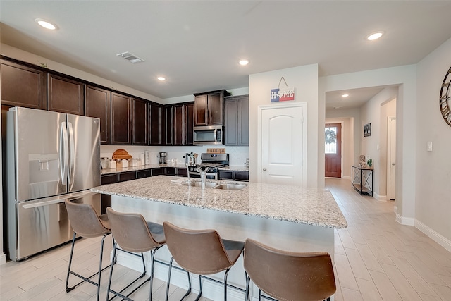 kitchen featuring dark brown cabinetry, a kitchen island with sink, stainless steel appliances, and light stone counters