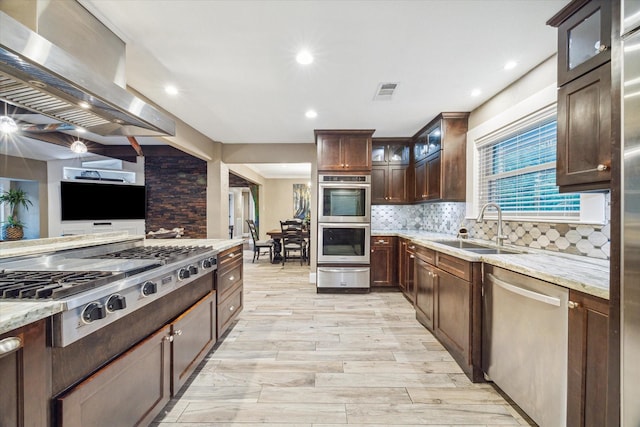 kitchen with island range hood, glass insert cabinets, appliances with stainless steel finishes, light stone countertops, and a sink