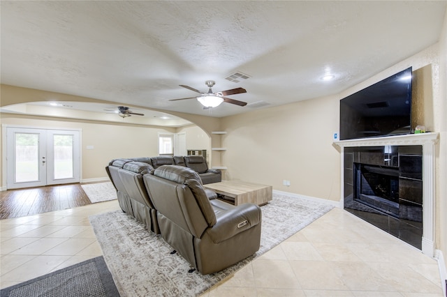 living room featuring a tile fireplace, light wood-type flooring, a textured ceiling, ceiling fan, and french doors