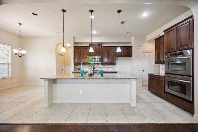 kitchen with an island with sink, appliances with stainless steel finishes, light tile patterned flooring, light stone countertops, and pendant lighting