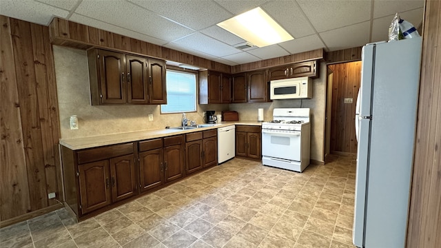 kitchen with dark brown cabinets, sink, a paneled ceiling, wooden walls, and white appliances