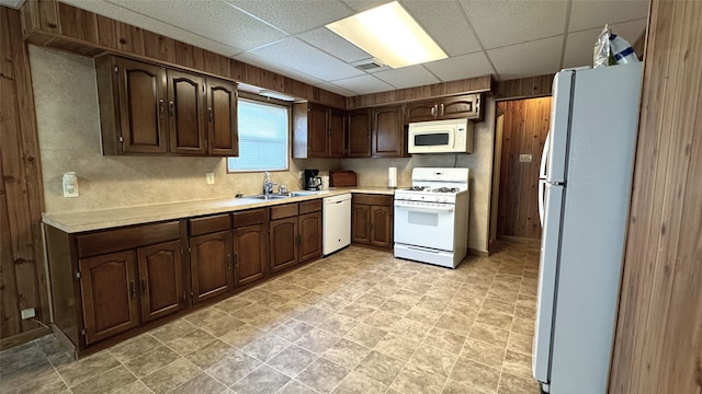 kitchen featuring dark brown cabinetry, sink, white appliances, wooden walls, and a drop ceiling