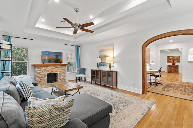 living room featuring light hardwood / wood-style floors, a stone fireplace, ceiling fan, and a tray ceiling