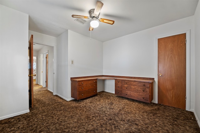 empty room with built in desk, dark colored carpet, and ceiling fan