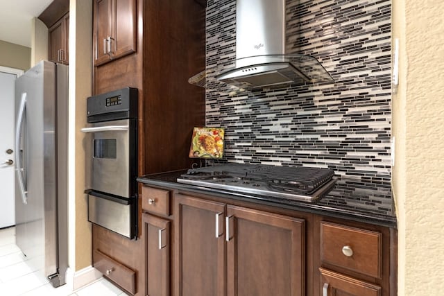 kitchen with backsplash, wall chimney range hood, dark stone countertops, light tile patterned flooring, and stainless steel appliances