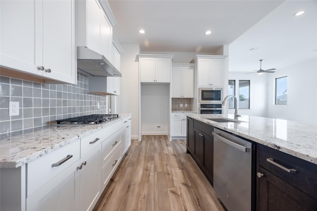 kitchen with wall chimney range hood, light wood-type flooring, appliances with stainless steel finishes, white cabinetry, and a sink