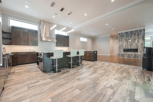 kitchen featuring dark brown cabinetry, a kitchen bar, a kitchen island, and wall chimney range hood