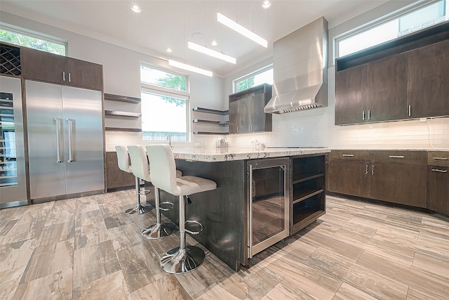 kitchen featuring dark brown cabinets, beverage cooler, wall chimney exhaust hood, and decorative backsplash