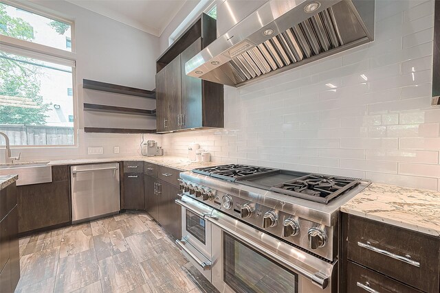 kitchen with dark brown cabinets, light stone counters, wall chimney range hood, decorative backsplash, and stainless steel appliances