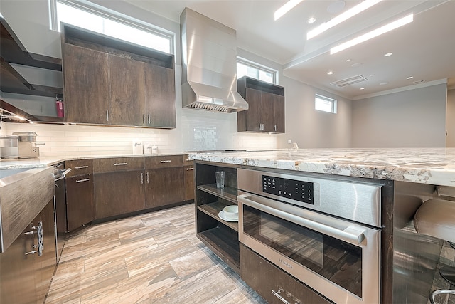 kitchen featuring dark brown cabinetry, oven, wall chimney range hood, and a healthy amount of sunlight