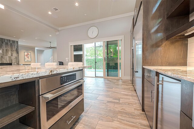 kitchen featuring light stone counters, oven, dark brown cabinetry, crown molding, and ceiling fan