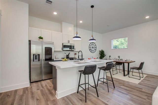 kitchen with white cabinets, an island with sink, pendant lighting, light hardwood / wood-style flooring, and stainless steel appliances