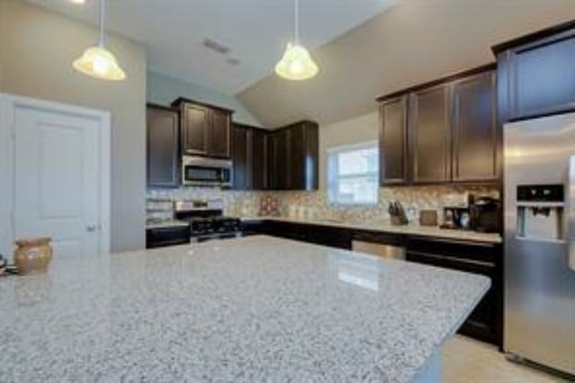 kitchen with lofted ceiling, dark brown cabinets, decorative light fixtures, stainless steel appliances, and light stone countertops