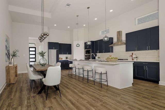 kitchen with wall chimney exhaust hood, hanging light fixtures, a high ceiling, dark hardwood / wood-style floors, and a center island with sink