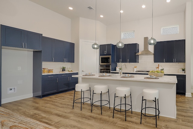 kitchen with a center island with sink, a towering ceiling, wall chimney range hood, and appliances with stainless steel finishes