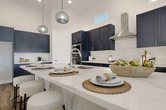 kitchen with wall chimney range hood, light stone counters, a towering ceiling, decorative light fixtures, and decorative backsplash