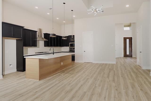 kitchen with a towering ceiling, light wood-type flooring, wall chimney range hood, pendant lighting, and a kitchen island with sink