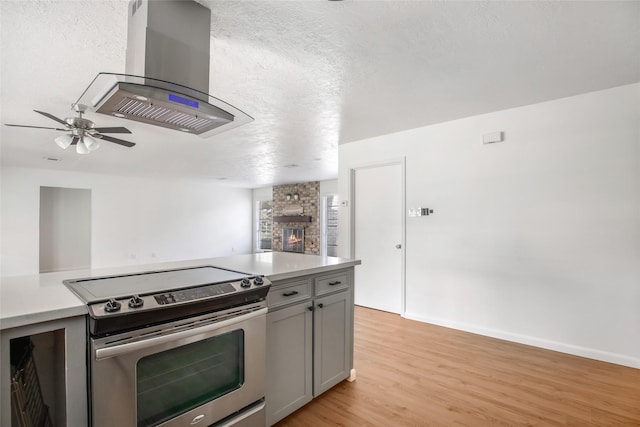 kitchen with stainless steel range with electric cooktop, light wood-type flooring, gray cabinets, island range hood, and a stone fireplace