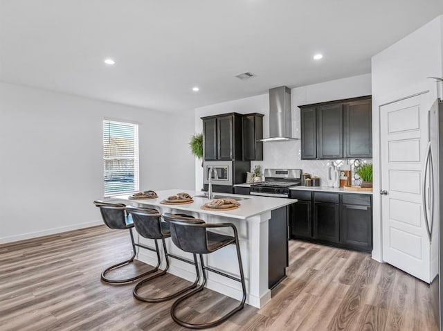 kitchen with light wood-type flooring, a kitchen island with sink, sink, wall chimney range hood, and stainless steel gas stove