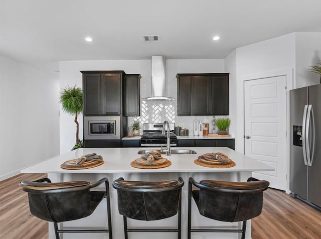 kitchen featuring an island with sink, tasteful backsplash, wood-type flooring, wall chimney range hood, and stainless steel appliances